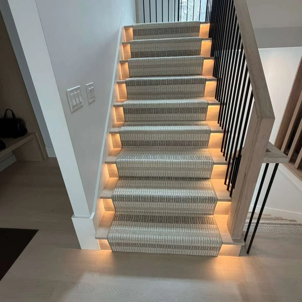 Wooden staircase with lighted treads and striped carpeting, black metal railing on one side, located in a modern home interior.