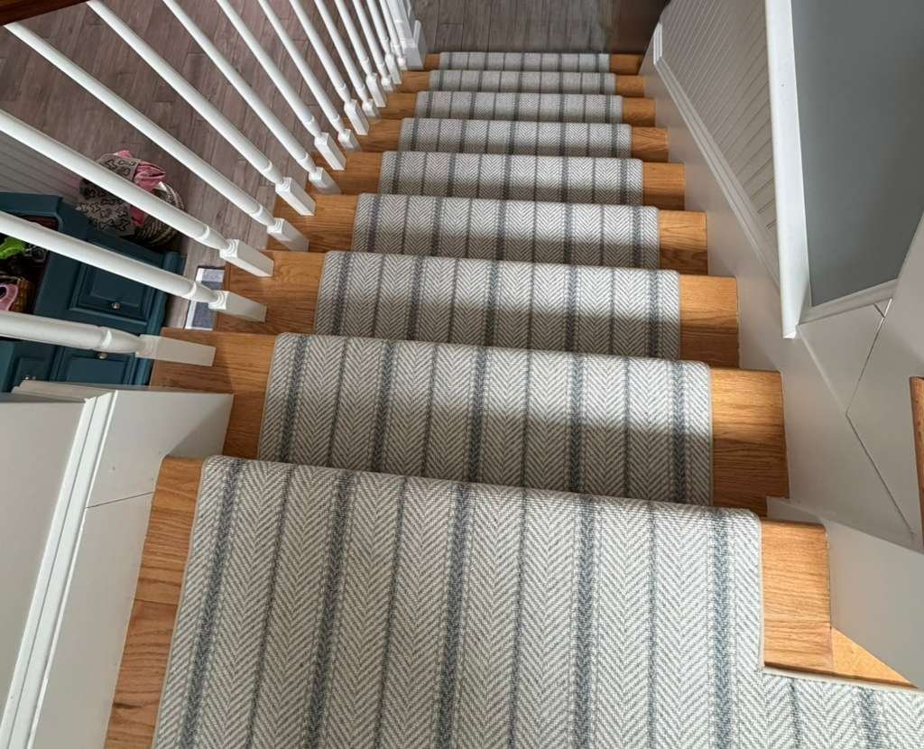 Wooden staircase with a gray and white striped carpet runner, white spindles, and part of a blue table and decorative lamp visible on the side.