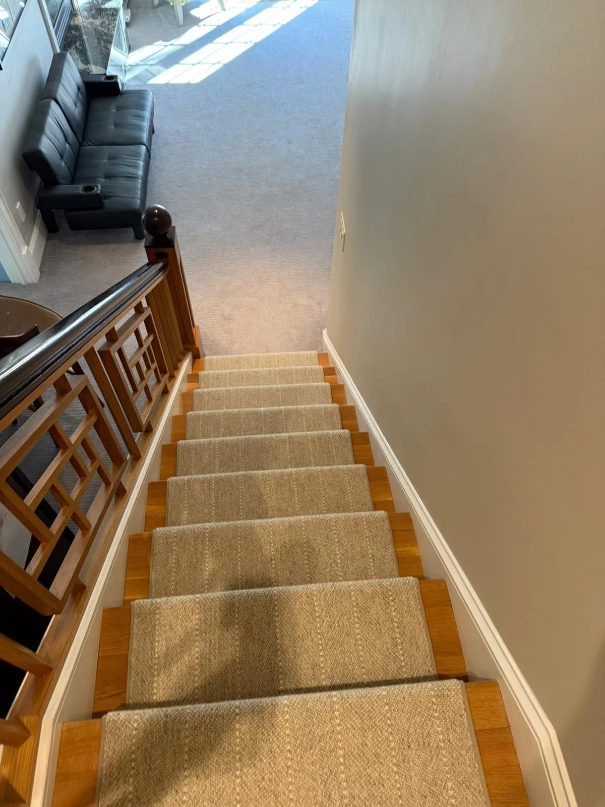 Top view of a staircase with beige carpeted steps, wooden trim, and a black handrail, leading to a carpeted lower level with a black sofa and a window area in the background.