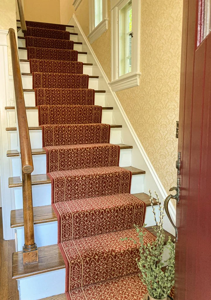 Interior view of a staircase with a red and gold patterned carpet runner, wooden handrail on the left side, and beige wallpaper with a floral pattern on the wall. There are three white-framed windows providing natural light.