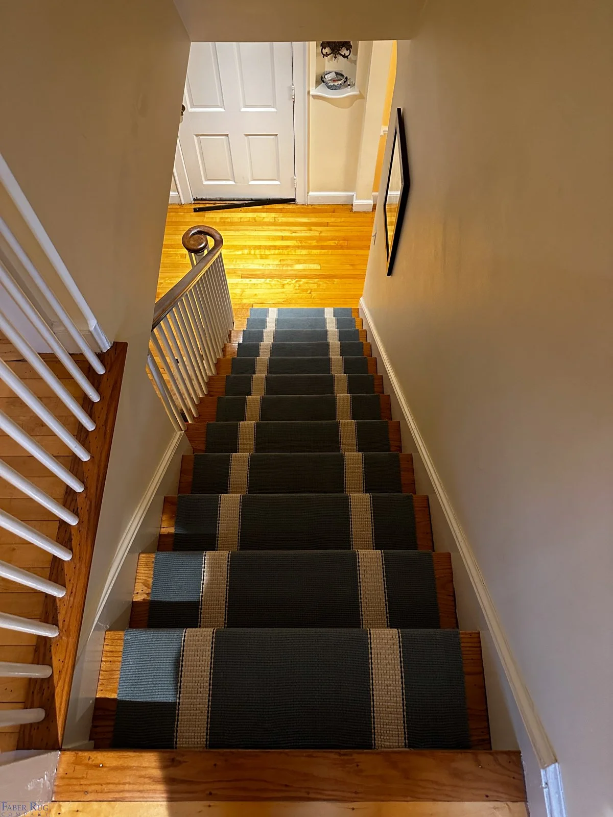 View of a staircase inside a house with a dark rectangular rug runner, beige walls, and a small hallway at the bottom leading to a closed white door.