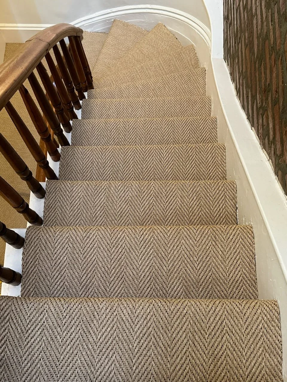 A view of a staircase with a beige herringbone-patterned carpet runner, a wooden handrail, white wall molding, and exposed brick on the side wall.
