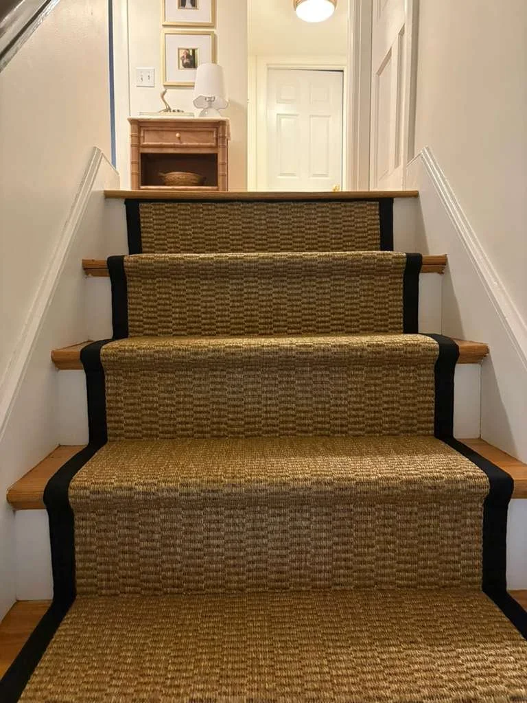 Interior view of staircase with beige sisal and black wide binding runner rug leading up to a hallway, with a wooden table and lamp on a side table at the top of the stairs.