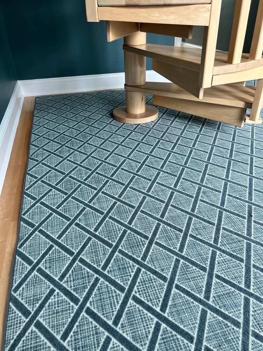 Close-up of a geometric patterned rug on a hardwood floor next to a dark teal wall. Part of a wooden staircase with a round support post is visible in the corner.