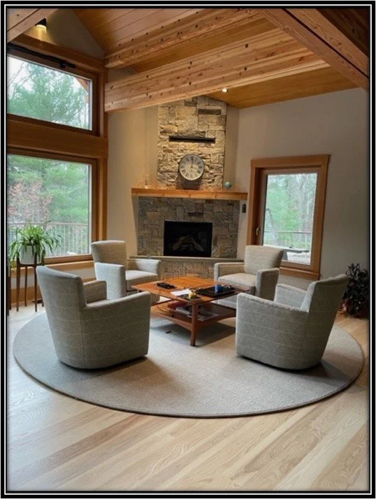 Living room with a stone fireplace, wooden ceiling, large windows, and a round rug with four beige armchairs and a wooden coffee table.