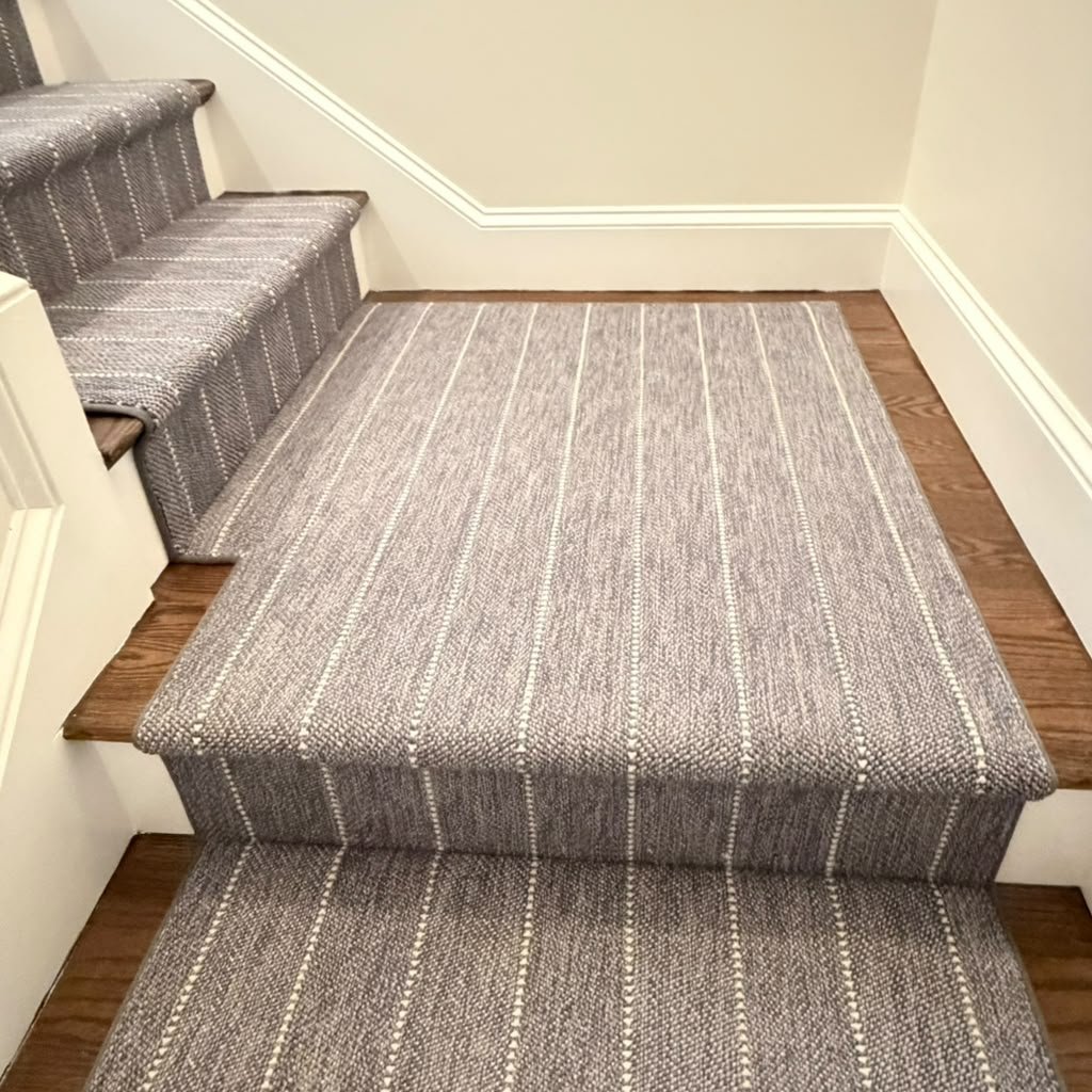 A staircase with a beige and gray striped carpeted runner, and a small landing area with matching carpet, next to hardwood flooring and white walls with white trim.