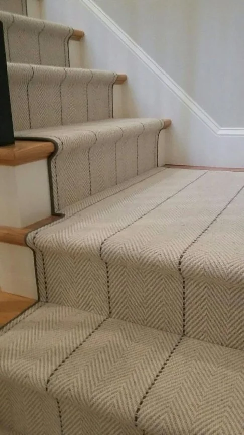 Carpeted staircase with beige and brown diagonal striped pattern, with wooden edges on some steps and white walls.