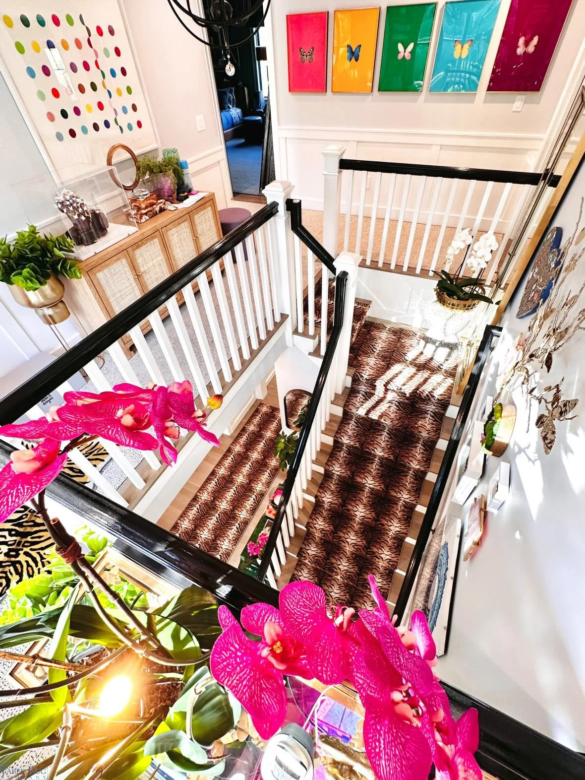 A view of a staircase landing from above, decorated with pink orchids in the foreground, framed butterfly artwork on the walls, a living room visible through the doorway, colorful framed butterflies on the wall, and a runner rug on the staircase.