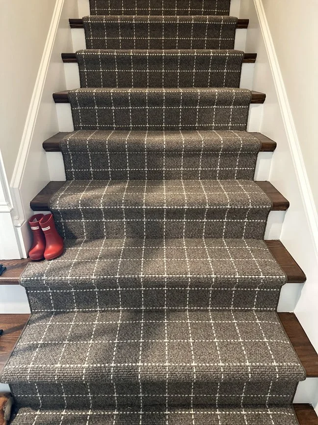 Carpeted staircase with dark wood trim on the steps and white walls on either side. There is a pair of red rain boots on the bottom step.