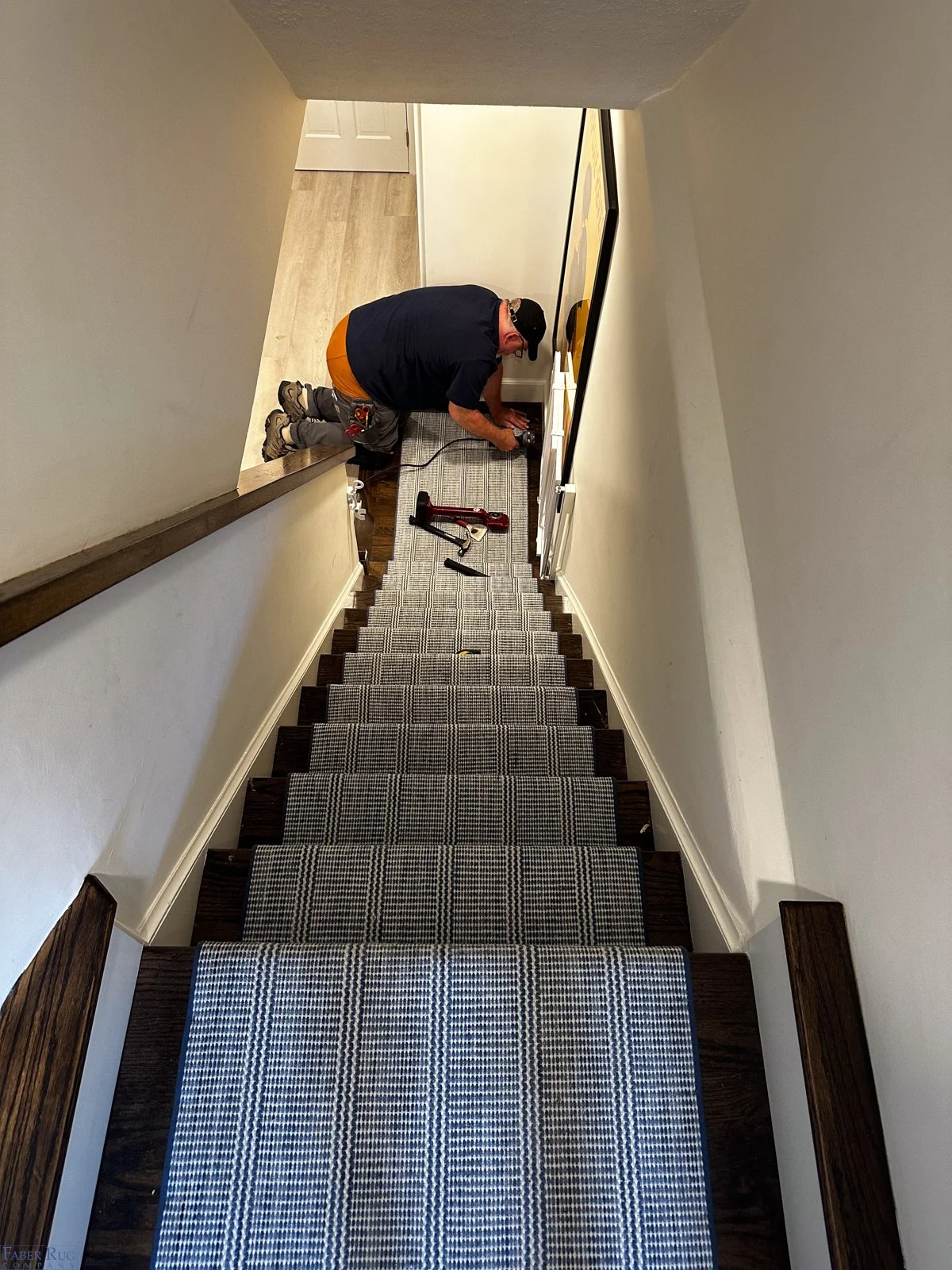A man kneeling on a staircase, using a power drill on the wall near the ceiling. Tools and a power tool are on the stairs.