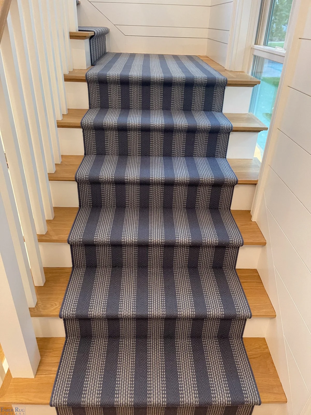 Wooden staircase with a striped carpet runner in shades of blue and beige, with natural light from a window on the right side.