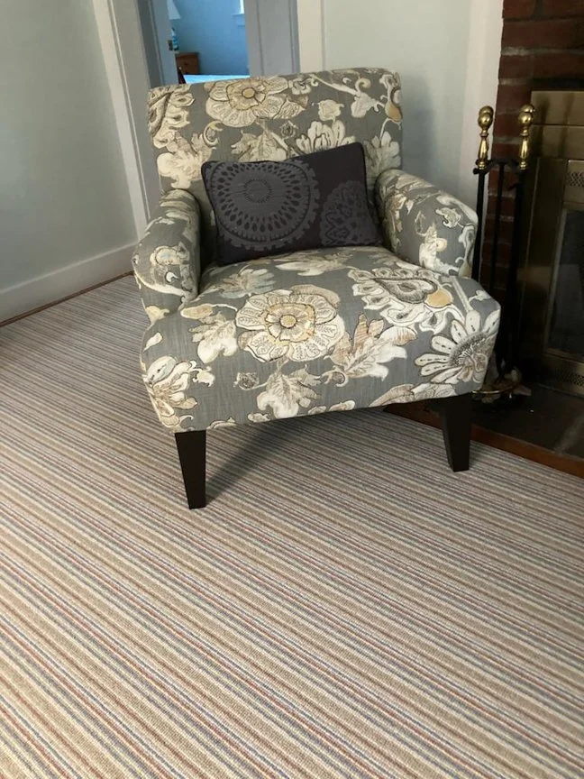 A floral patterned armchair with dark wooden legs, placed on a striped multicolored carpet, next to a fireplace with brass and black tools, in a room with light-colored walls.