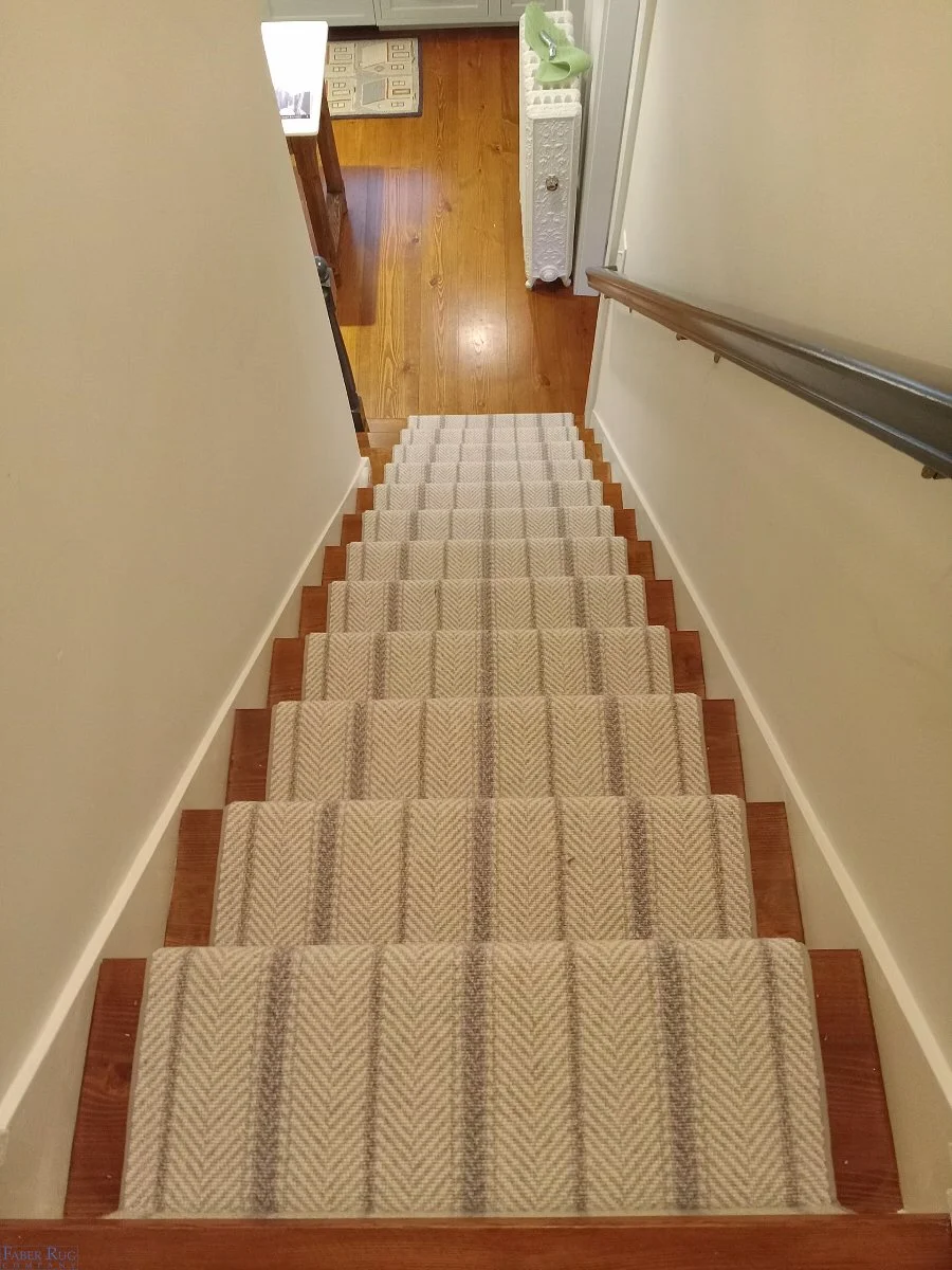 Top of a carpeted staircase showing beige zigzag-patterned carpet runner and wooden steps, leading down to a hardwood floor with a small beige and green rug, a white decorative end table, and a green lamp.