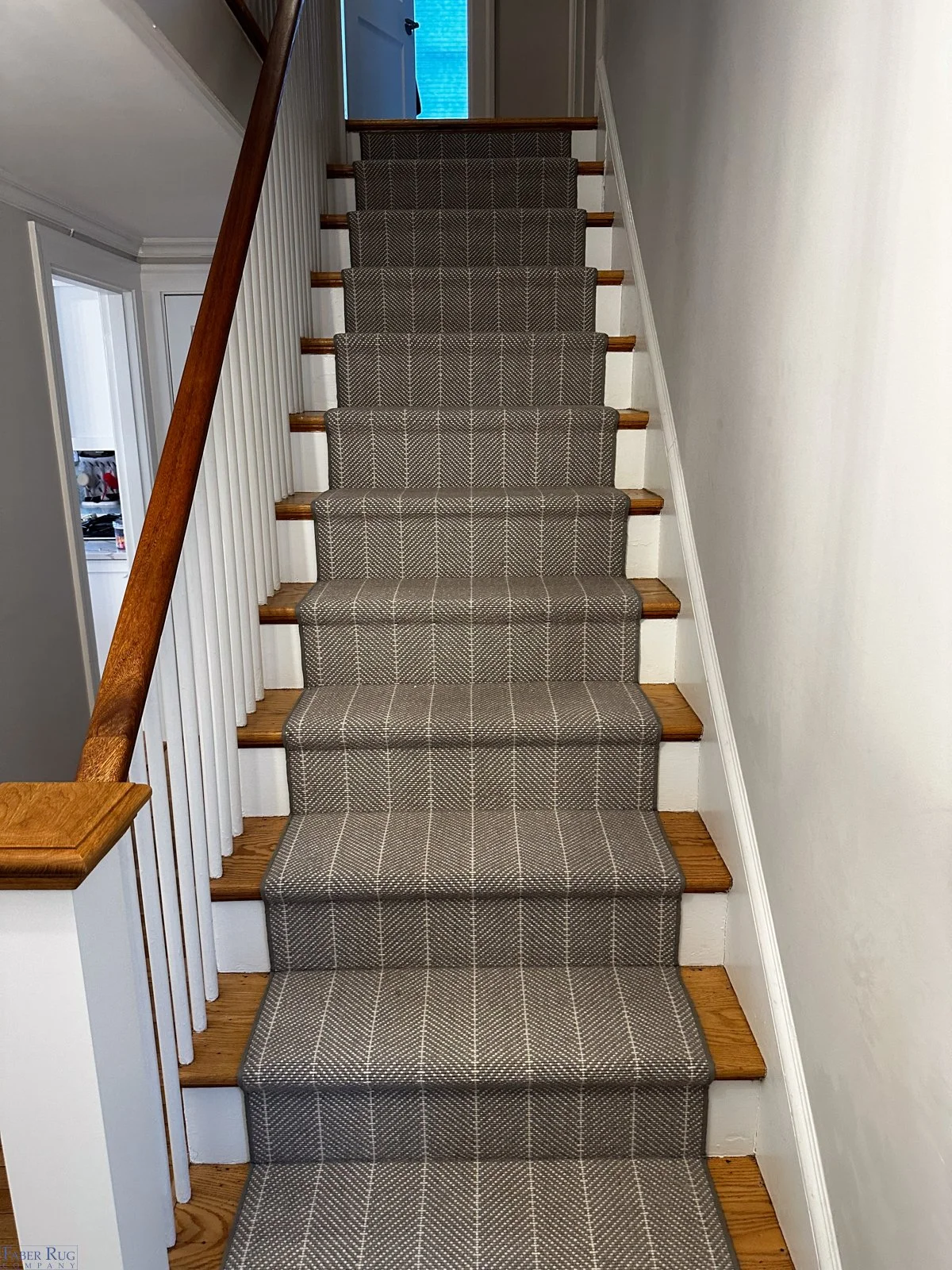 Carpeted staircase with a checkered pattern and wooden steps, white wall on right, white railing and wall on left, with a door at top showing a blue exterior.