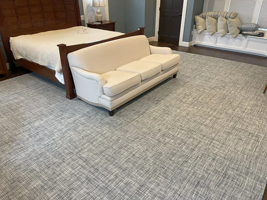 A bedroom with a large bed, beige sofa, window seat with pillows, and a carpeted floor.