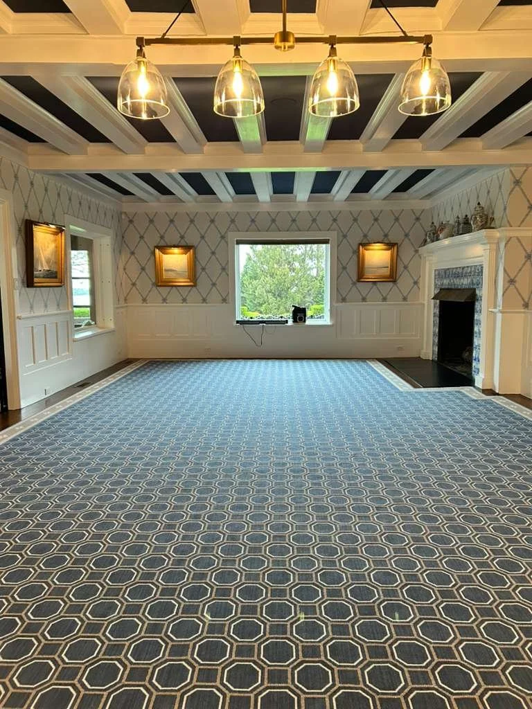 Empty living room with patterned blue carpet, white paneled walls, three hanging pendant lights, a large window, and a fireplace