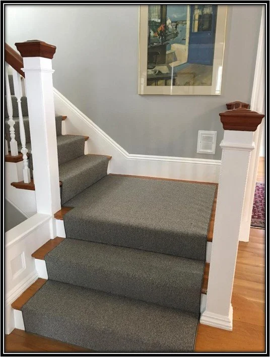A staircase with wooden steps and white railings, carpeted with dark gray runner, leading to an upper floor. A framed artwork hangs on the gray wall above the landing, and a small vent is visible on the wall. The flooring at the bottom is hardwood.