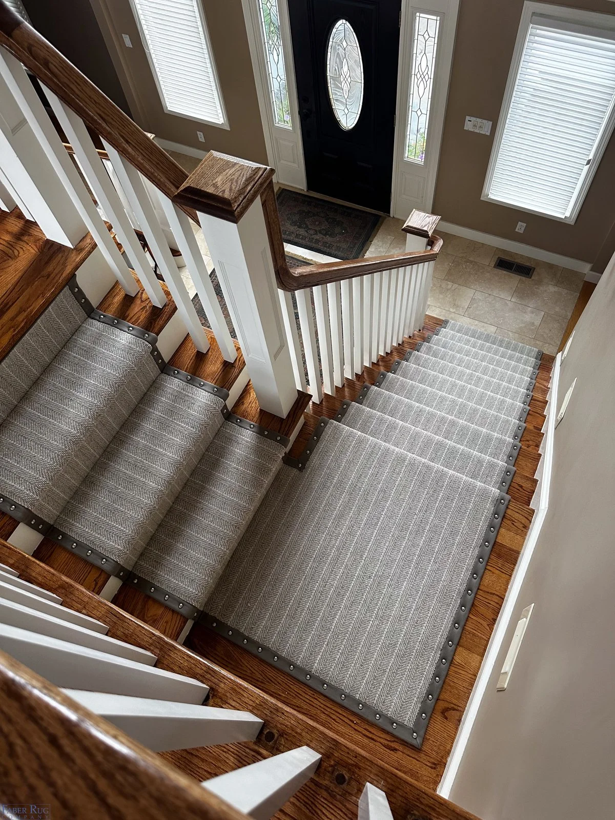 View from the top of a staircase looking down at an entrance door with two windows, a tiled floor, and a shoe mat, with carpeted stairs and wooden handrails.