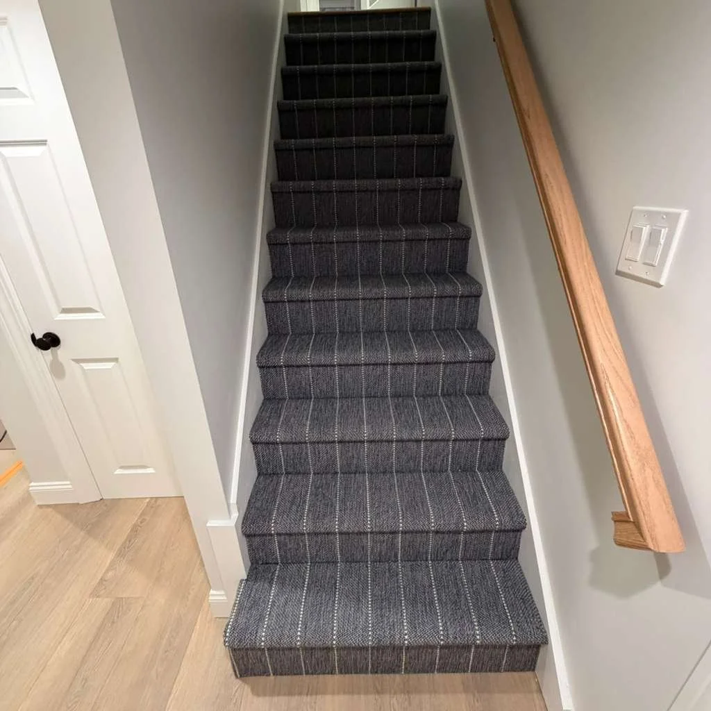 Carpeted stairs with a dark gray, plaid pattern, leading upward, flanked by white walls and a wooden handrail on the right.