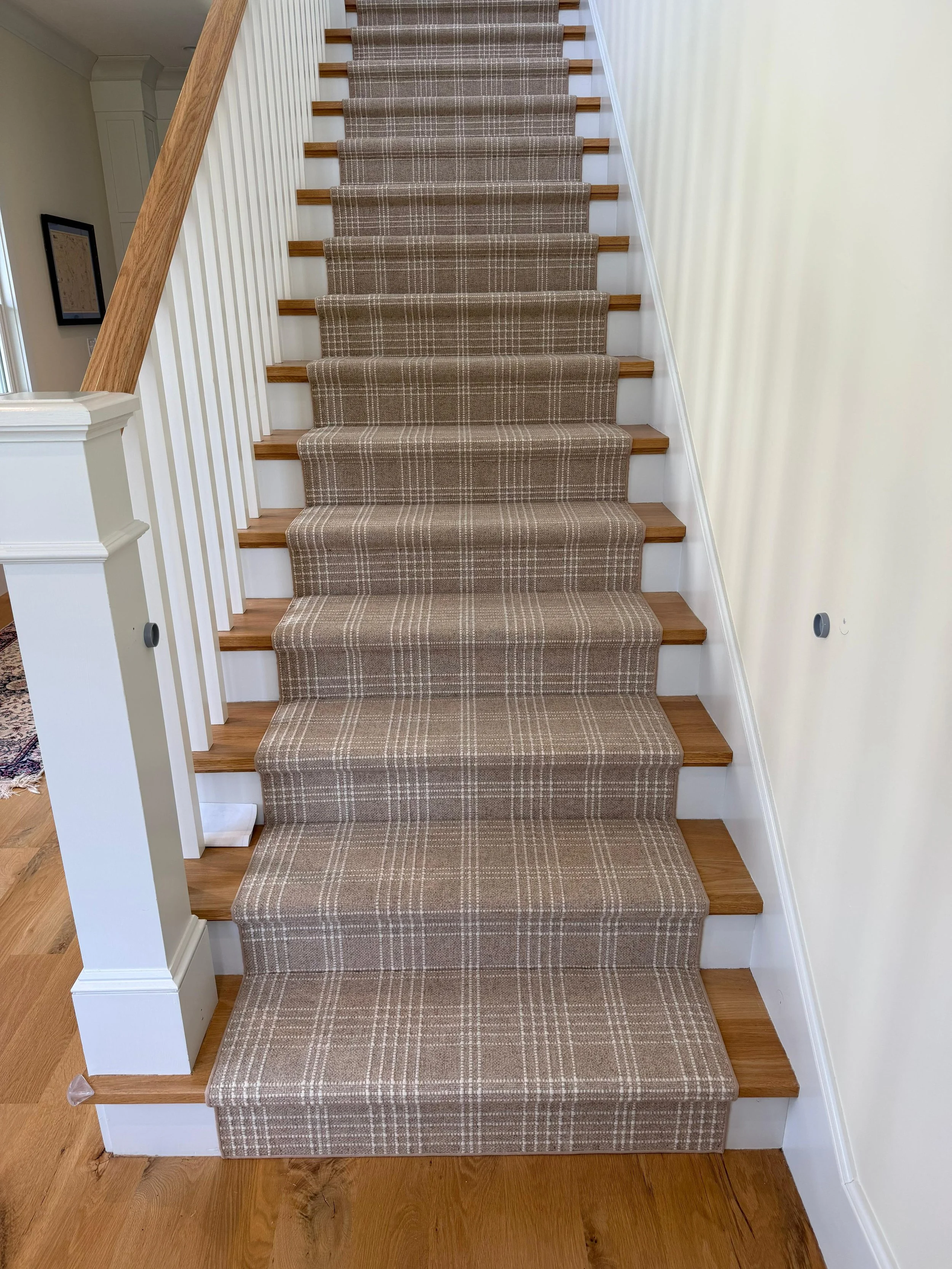 A staircase with a patterned runner carpet, wooden treads, white risers, and a white handrail on the left side, with a light-colored wall on the right side.