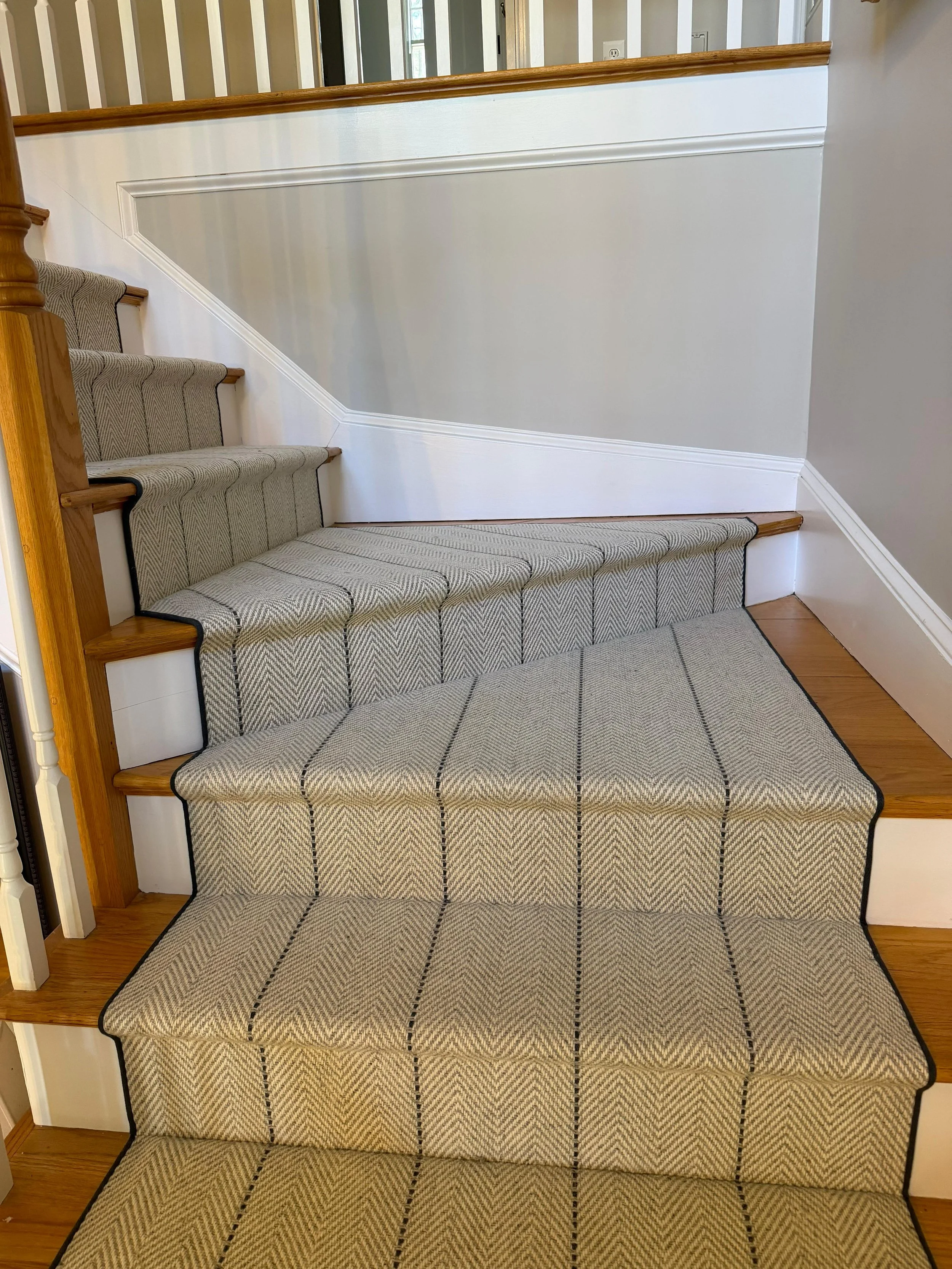 A staircase with patterned carpet and a white half wall with a wooden handrail at the top, with visible doorways and windows in the background.