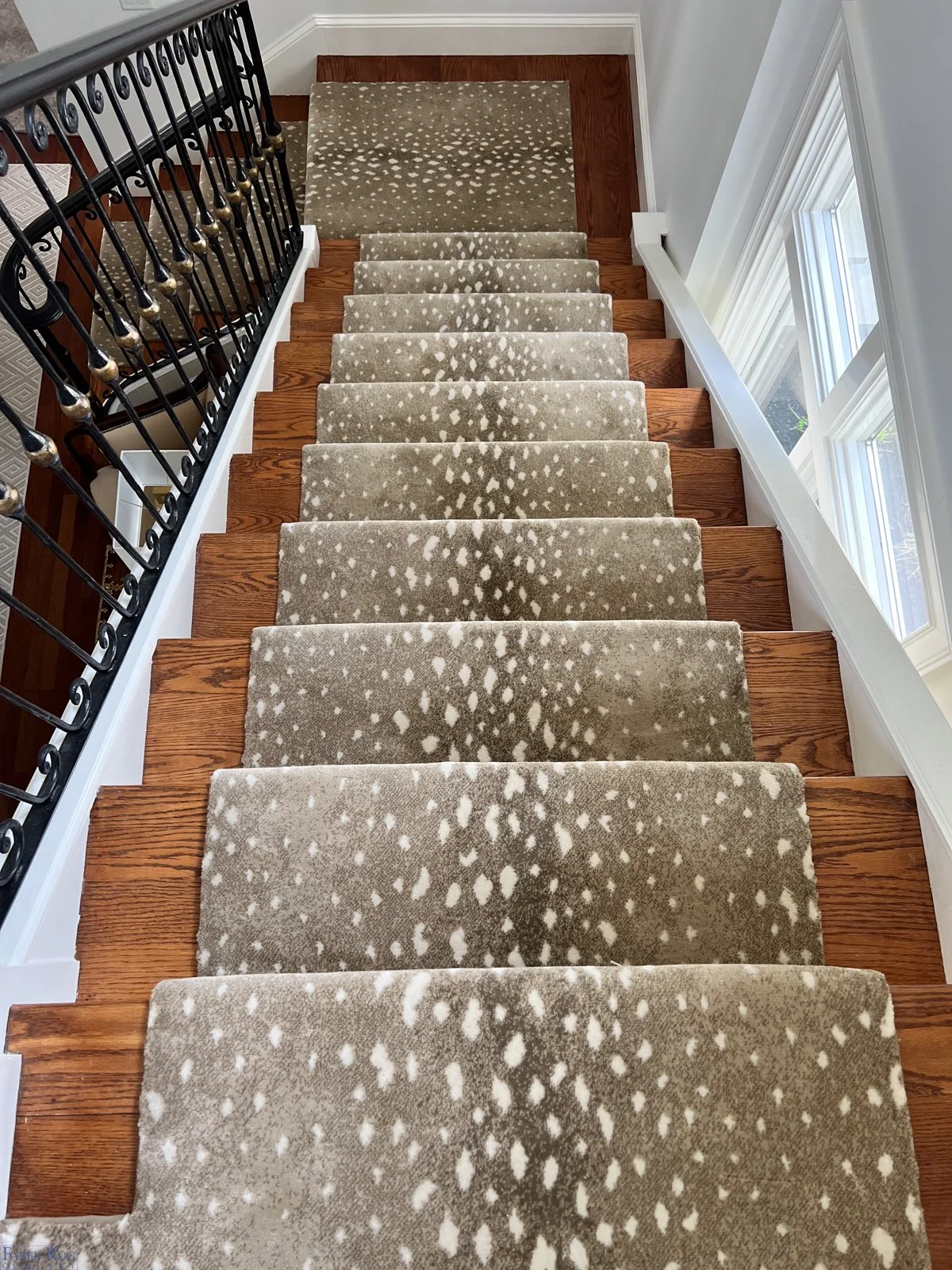 Top-down view of a staircase with a patterned beige and white runner, wooden steps, white trim, and a large window on the right, with a black wrought iron railing on the left.