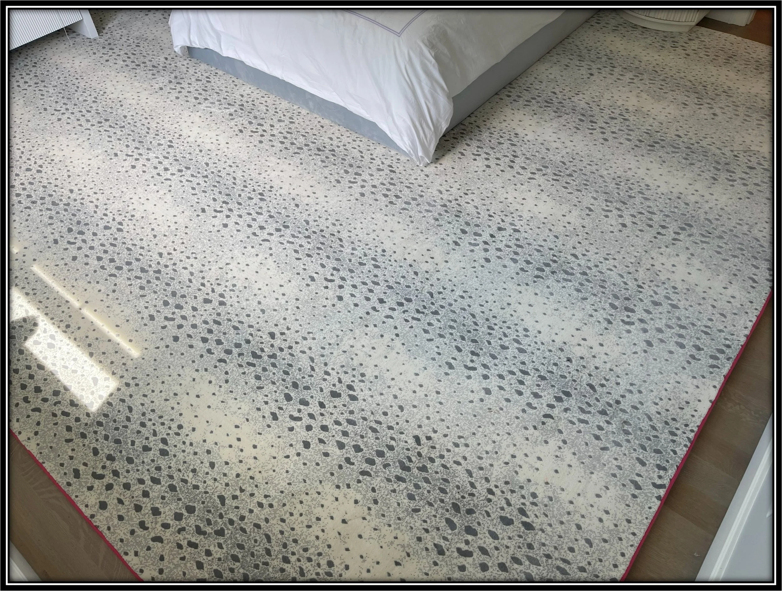 Close-up of a speckled white and gray rug underneath a bed with white bedding.