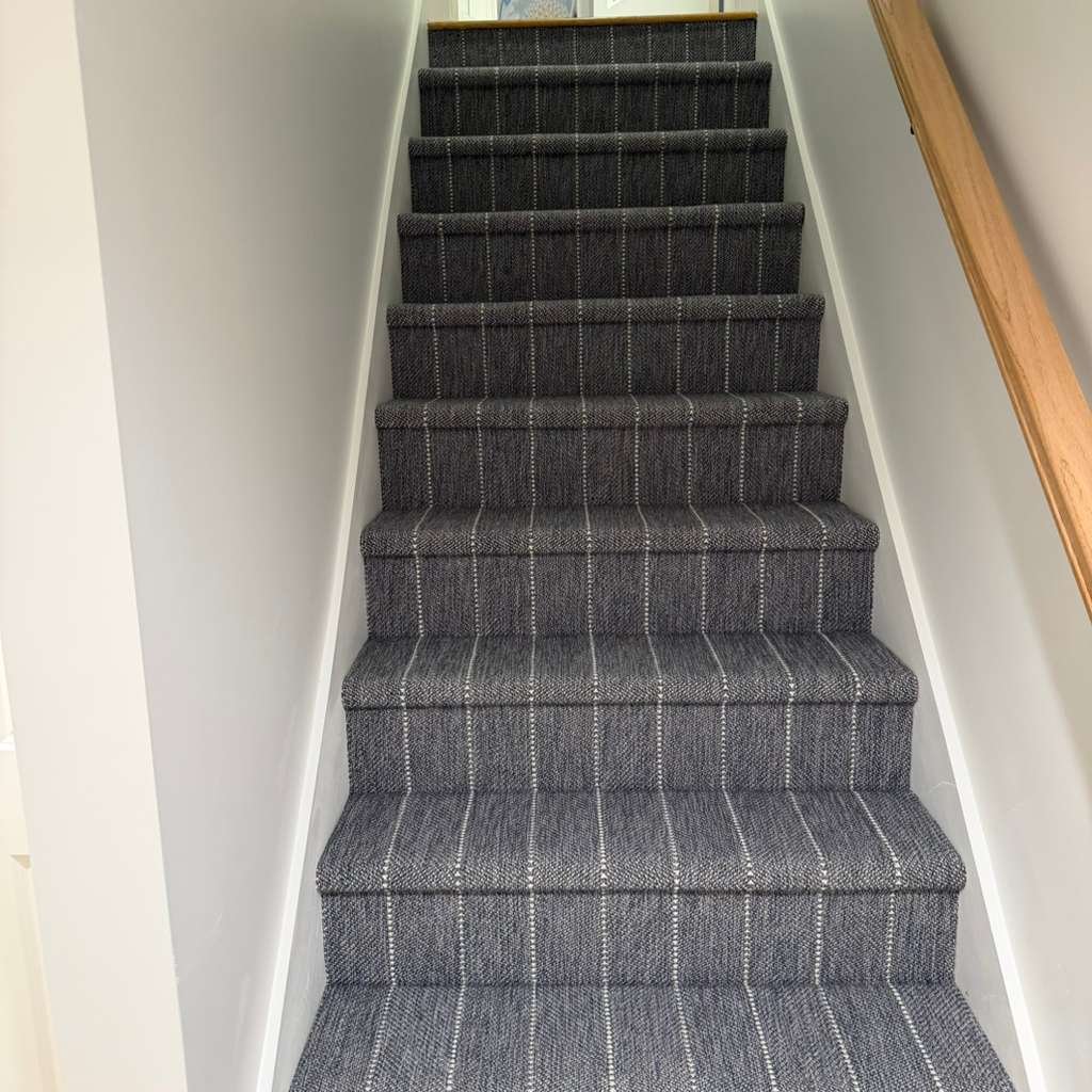 Indoor staircase with gray plaid carpeted steps and white walls on either side, with a wooden handrail on the right.