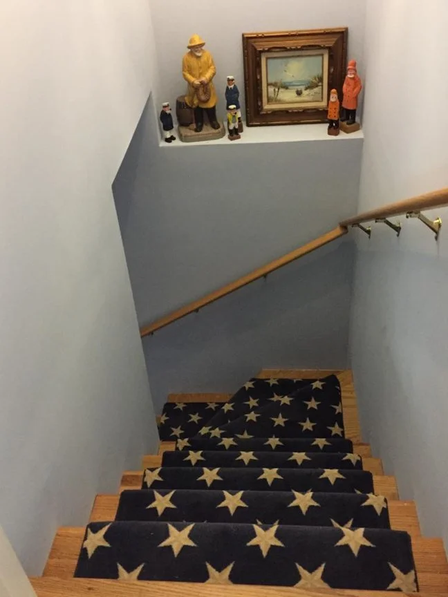 View from the top of a staircase with a black rug featuring a cream star pattern. At the landing, a display shelf holds a framed painting of a seascape and several small decorative figurines. The stairs descend downward, with a wood handrail on the r