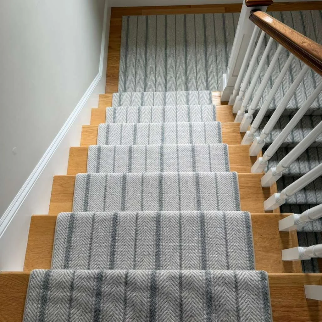 View of a staircase from above, with gray and white striped carpeted steps, wooden edges, white spindles, and a wooden handrail. A porch and part of a wall are visible at the top.