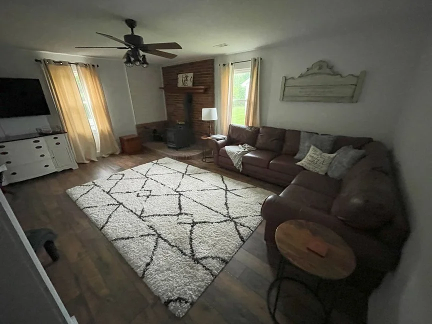 Living room with a large brown sectional sofa, a white shag rug with black lines, a fireplace, a window with beige curtains, a wall-mounted TV, and a ceiling fan.