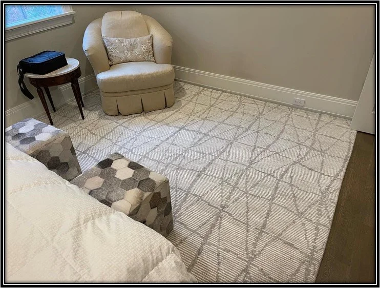 A cozy corner of a bedroom with a beige armchair, a small wooden side table with a black bag on it, a patterned ottoman, and a large light-colored area rug with a geometric design.