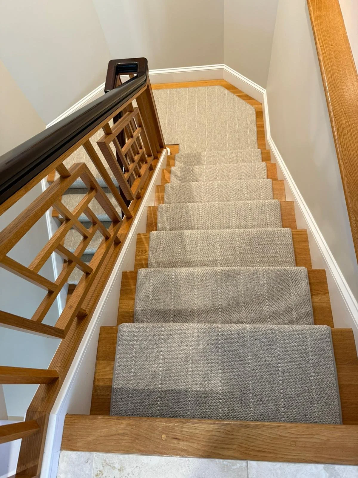 Overhead view of a staircase with beige carpeted steps, wooden handrail, and a small landing at the top with a carpeted area and wooden trim.