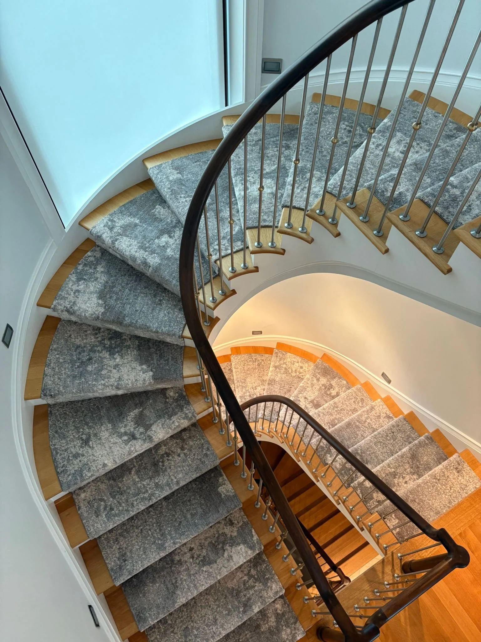 A winding staircase with a carpet runner, viewed from above, with a curved wooden handrail and metal balusters.