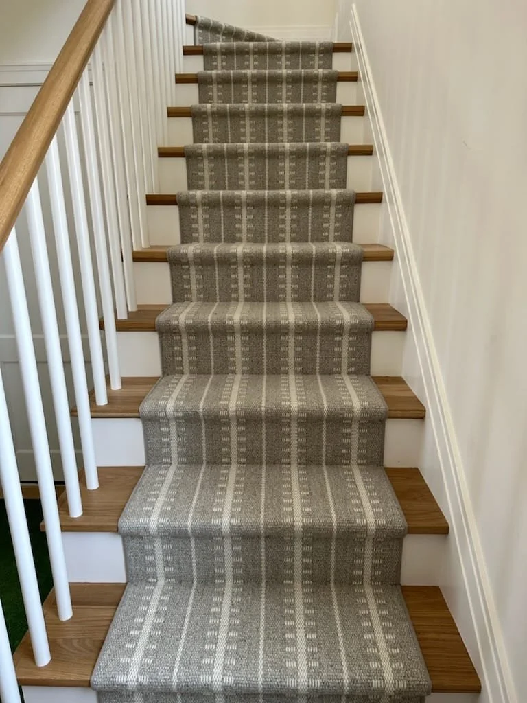 Carpeted staircase with gray patterned runner, wooden steps, white walls, white spindle railing on the left, and wooden handrail on the left side.