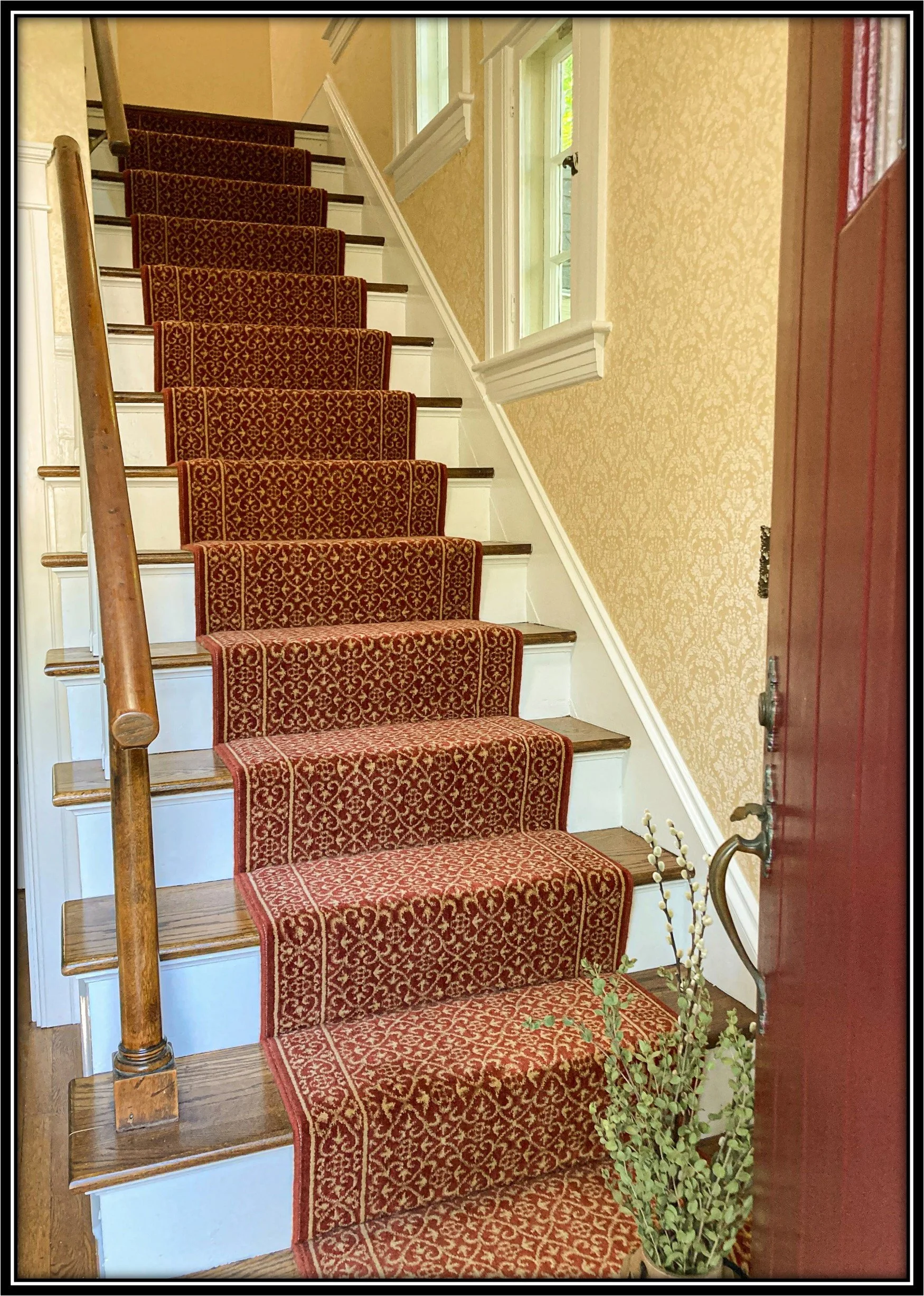 Interior view of a staircase with a red ornate patterned carpet runner, white risers, dark wooden treads, wooden handrail, beige wallpaper with a subtle design, a window with white trim, and a potted plant at the bottom of the stairs.