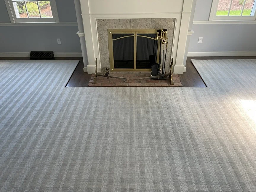 A living room with a fireplace, framed by a white mantel and a brick hearth, flanked by two windows letting in natural light, with beige striped carpet covering the floor.