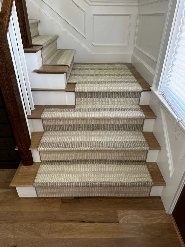 A staircase with carpeted steps in a house interior, with beige and brown striped carpet runners on some steps, and wooden edges around others. There is white wainscoting on the wall and a window with blinds to the right.