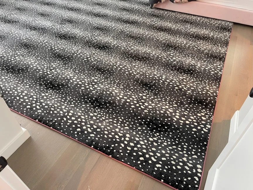 Close-up of a black area rug with small white and beige dot patterns near a bed and floor.
