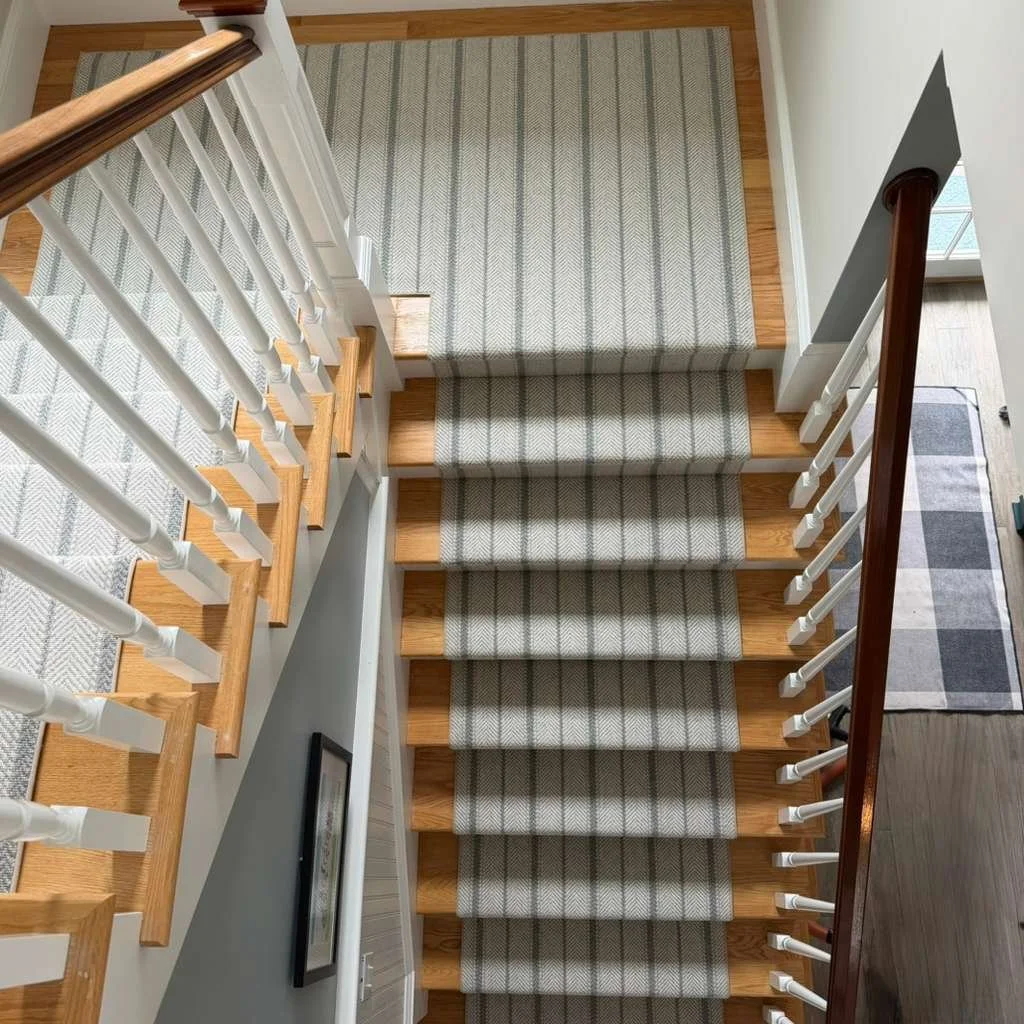 View of a staircase from above, showing steps with beige plaid carpet runners, white spindles, and wooden handrails, with a patterned rug at the bottom and a wall with a framed picture on the side.