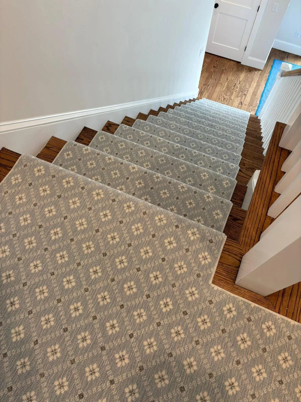Overhead view of a staircase with a floral-patterned carpet runner down the middle, brown wooden stairs, white walls, and a blue doormat at the bottom near a white door.