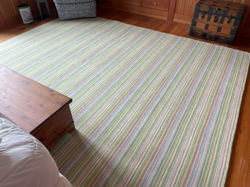 Room with a multicolored striped area rug on wooden floor, part of a bed, a wooden dresser, and a decorative chest.