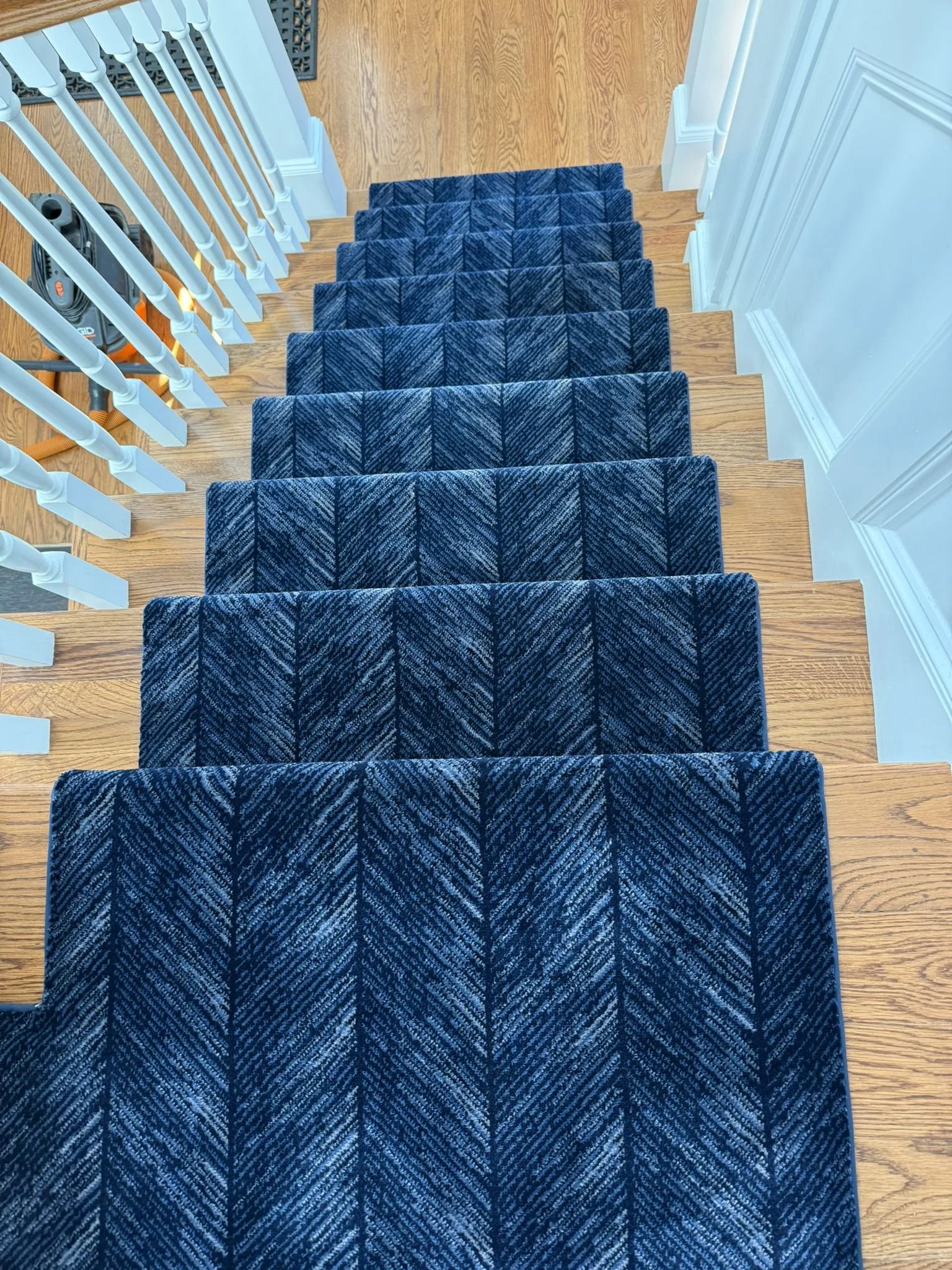 Top-down view of a staircase with a blue patterned carpet runner, wooden steps, white railings, and a vacuum cleaner to the left.