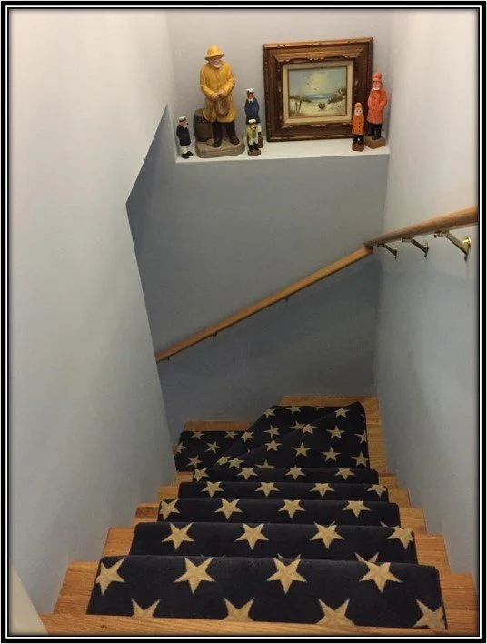 View of a staircase with a star-patterned carpet runner, leading down to a landing with a light blue wall decor shelf holding wooden figurines and a landscape painting.