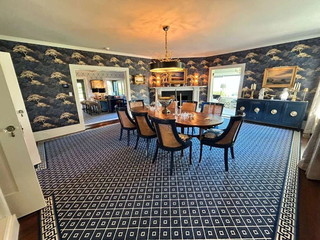 A dining room with a wooden table, eight matching chairs, a blue sideboard with candles, and a fireplace. The room has dark patterned wallpaper, a patterned carpet, and large windows.