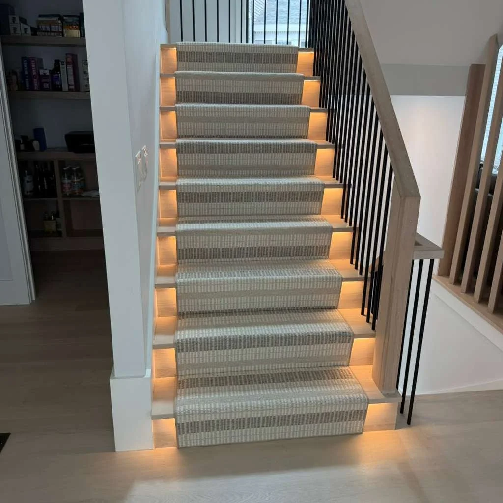 Interior view of a staircase with beige striped carpeted stairs, black metal railing on one side, and built-in lighting underneath each step, in a modern home.