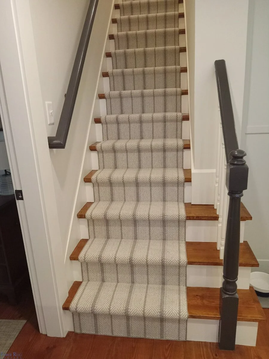 Indoor staircase with beige and brown plaid carpet runner, wooden treads, white risers, black handrails on both sides, and white walls.