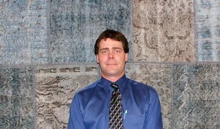Mike in a blue dress shirt and patterned tie standing in front of a textured wall background.