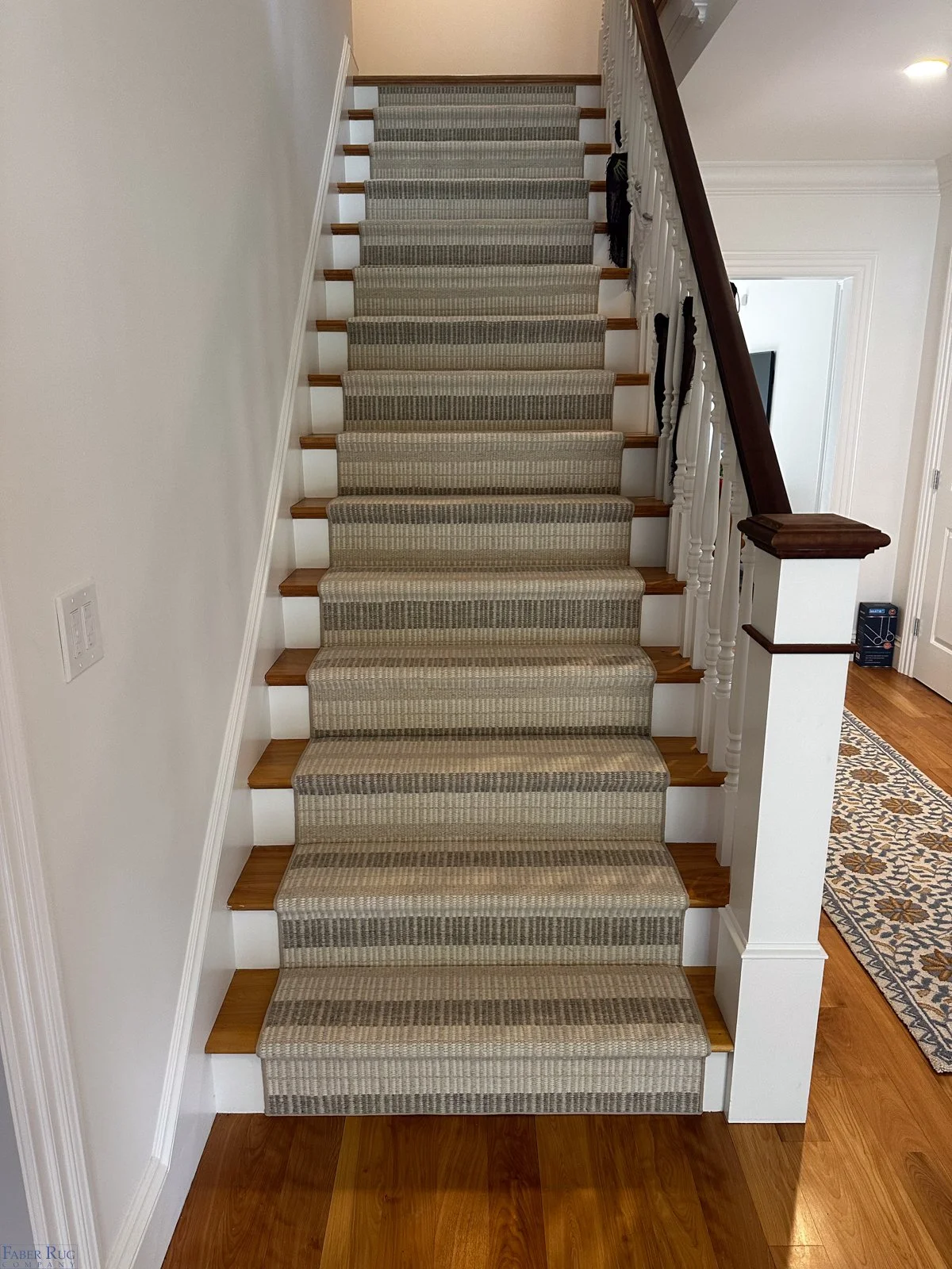 Carpeted staircase with white risers, wooden treads, and a white banister with decorative balusters. To the right, there is a dark wooden handrail and part of a wooden-newel post. The staircase is in a well-lit interior space with hardwood flooring a
