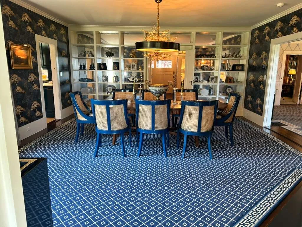 A formal dining room with a large wooden dining table, twelve chairs with blue frames and cane backs, an ornate gold chandelier, and built-in white bookshelves filled with decorative items, with dark floral wallpaper and a patterned blue and white ar